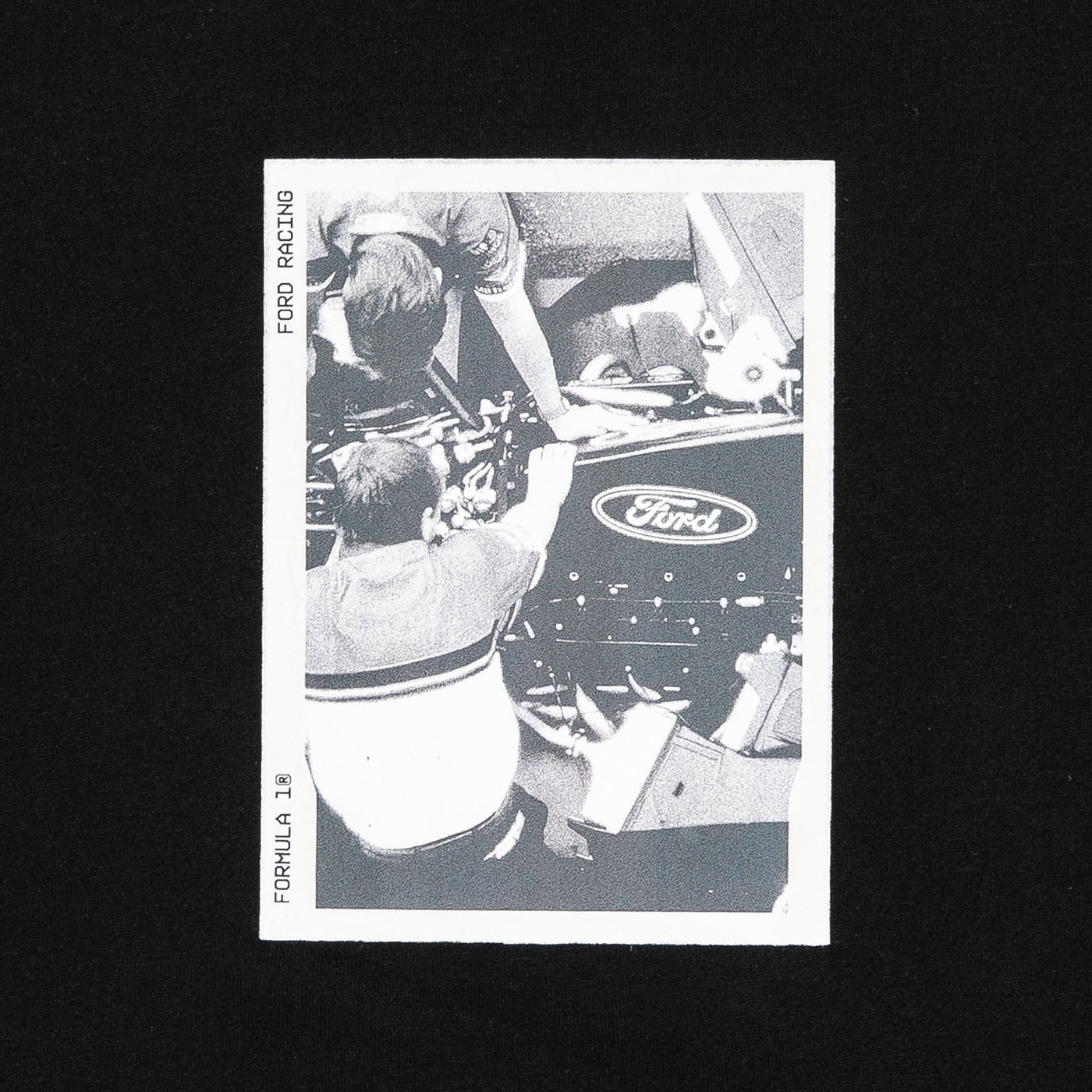 A vintage black and white Ford racing collectible card featuring a photo of mechanics working on a race car, displayed on a solid black background.