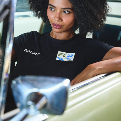 A woman with curly hair leans against the open door of a vintage car, wearing a black t-shirt with white 'Formula 1' branding and a 125th Anniversary patch.