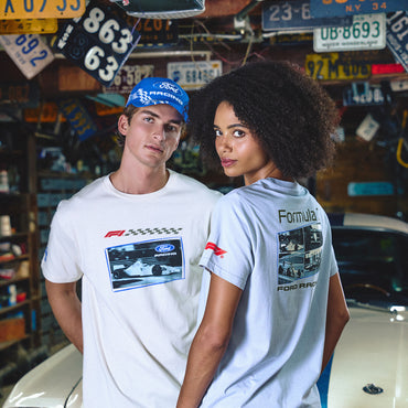 Two models wearing Ford Racing branded t-shirts in a garage setting filled with vintage license plates and a classic car. The male model wears a white crewneck shirt with a blue and white checkered F1 graphic, and the female model wears a light blue shirt showing the back with a Formula 1 Ford Racing graphic.