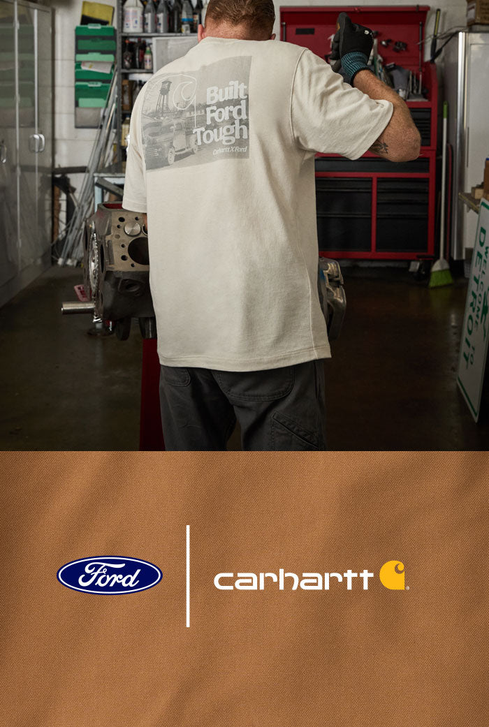 A man stands with his back to the camera in an auto repair shop, wearing a white T-shirt with a gray graphic on the back that reads 'Built Ford Tough' and dark pants. The bottom of the image features a brown graphic panel with the Ford logo and the Carhartt logo.