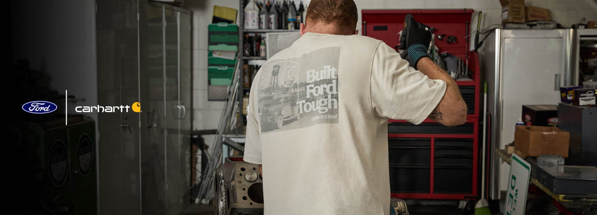 A man standing with his back to the camera in a garage, wearing a light beige Ford Tough T-shirt with a large graphic print on the back. The shirt features the text 'Built Ford Tough' and a gray graphic.