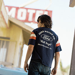 A man viewed from behind wearing a navy blue Ford short-sleeve t-shirt with orange and white stripes on the sleeves and a large white 'Ford' logo on the back, standing in front of a motel sign.