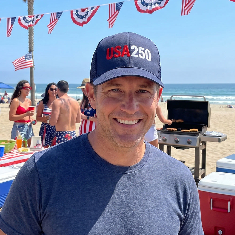 Man wearing a cap with 'USA 250' at a beach barbecue with people and flags in the background.