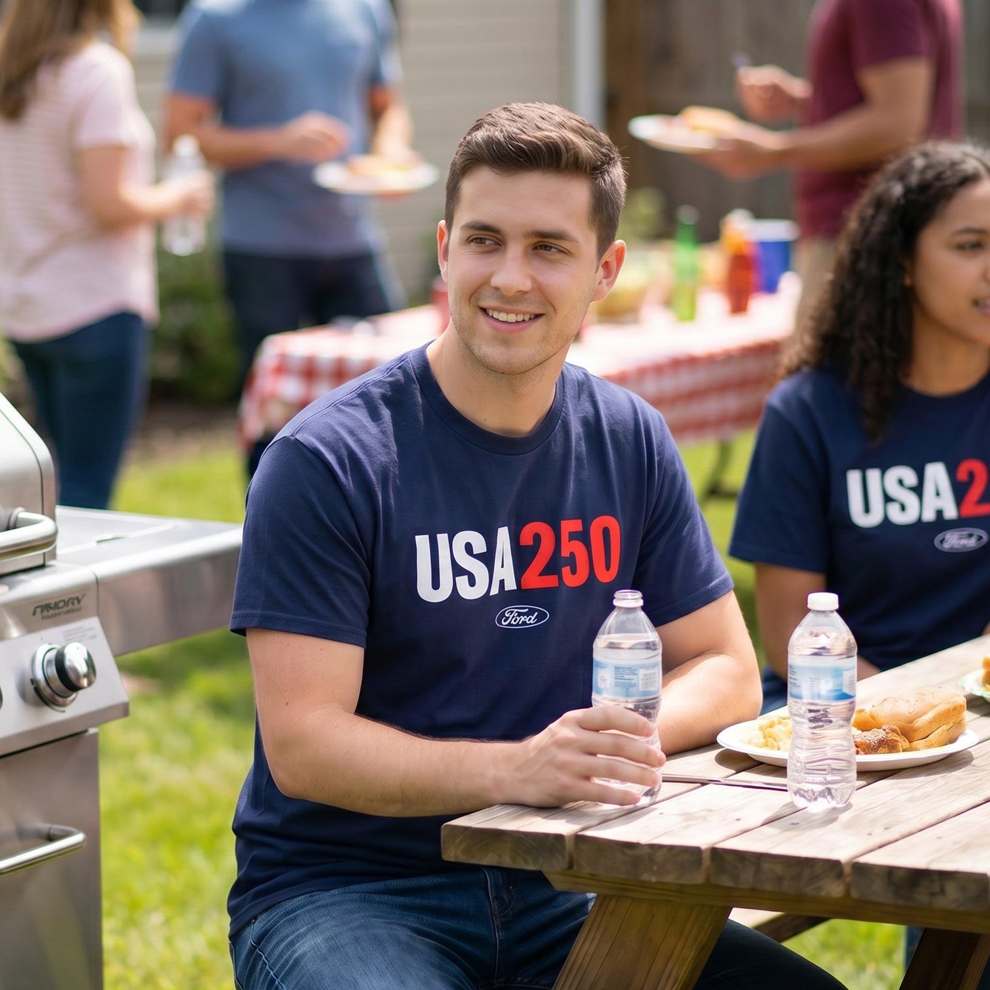 Man sitting at a picnic table with a 'USA 250' t-shirt, holding water bottles, with people and food in the background.