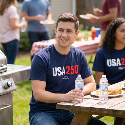 Man sitting at a picnic table with a 'USA 250' t-shirt, holding water bottles, with people and food in the background.