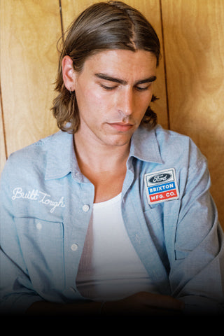 A man with long brown hair looking down, wearing a light blue Ford work shirt over a white undershirt, set against a warm wood-paneled background.