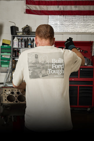 A man viewed from behind in a garage workshop, wearing a cream-colored t-shirt with a large black and white graphic on the back that reads 'Built Ford Tough'.