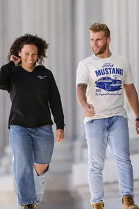 Two people walking side by side, one wearing a Ford Mustang Womens black sweatshirt and jeans, the other wearing a white t-shirt with a Ford Mustang graphic and jeans.
