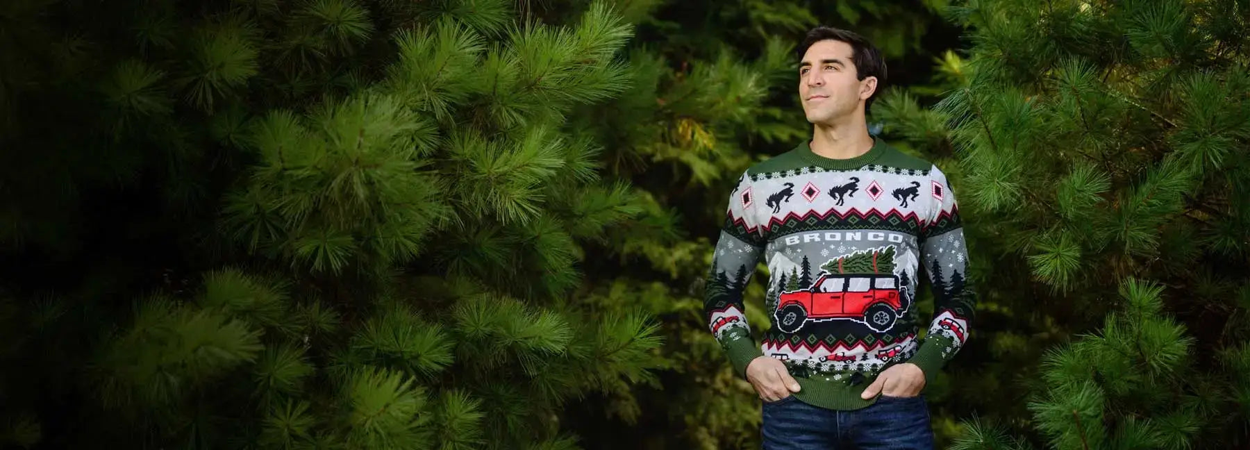 Man wearing a festive sweater with a train design, standing against a background of green trees.
