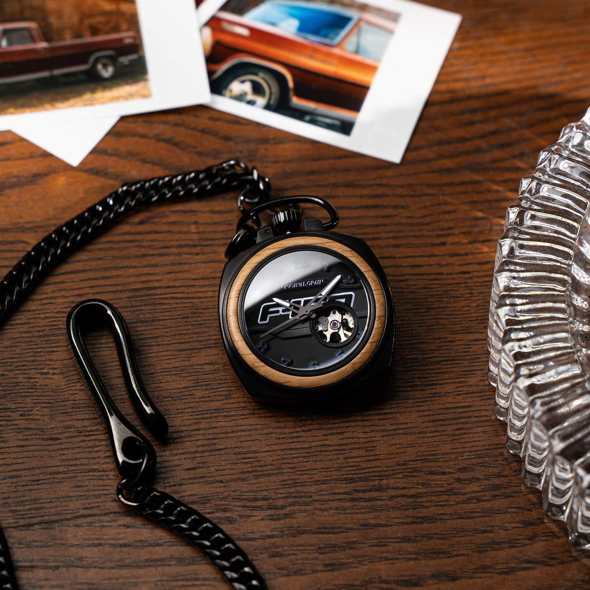 Pocket watch with wooden face on a wooden surface with photos and a glass object in the background