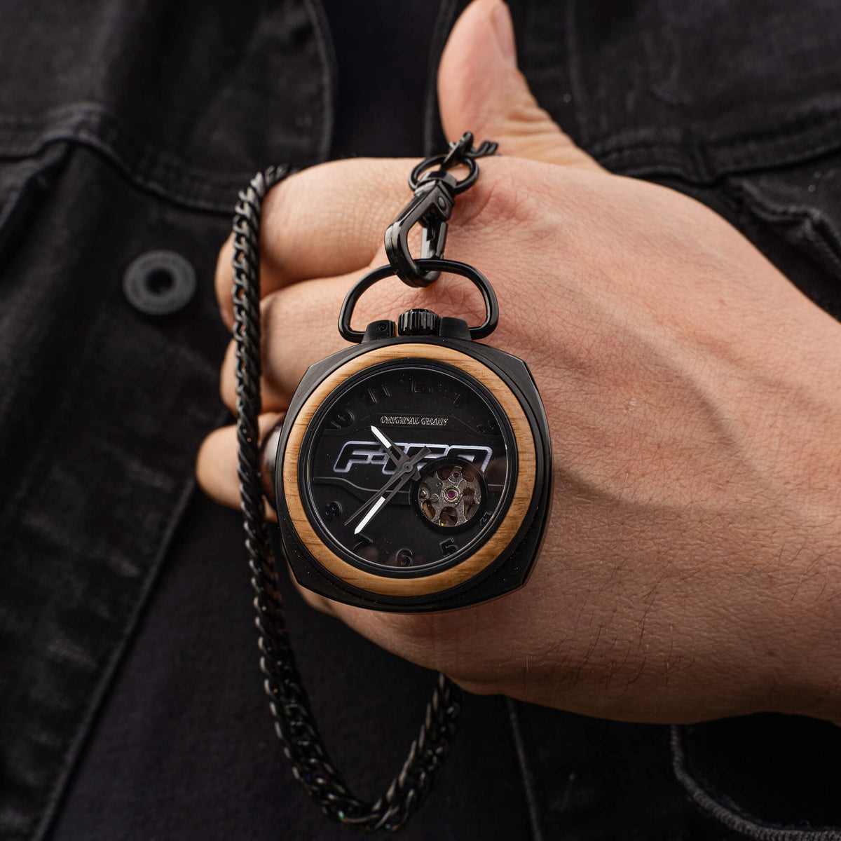 Hand holding a wooden and black F-150 pocket watch with a brand logo, against a dark background.